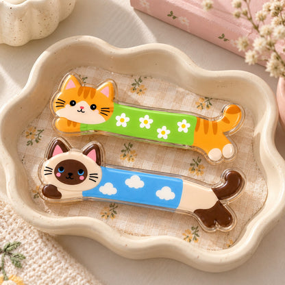 Kittens Day hair clips flat lay: orange tabby cat in a green daisy jumper and Siamese cat in a blue cloud jumper